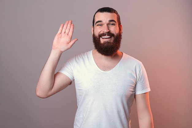 Giovane uomo barbuto amichevole sta mostrando ciao gesto alla fotocamera Studio girato su sfondo grigio