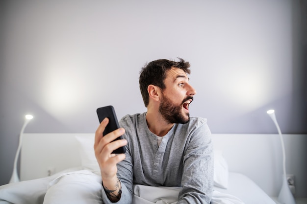 Giovane uomo barbuto bello che si siede nel letto in camera da letto e che tiene Smart Phone