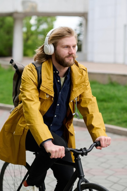 Giovane uomo barbuto che ascolta musica in cuffie wireless mentre guida una bicicletta lungo la strada