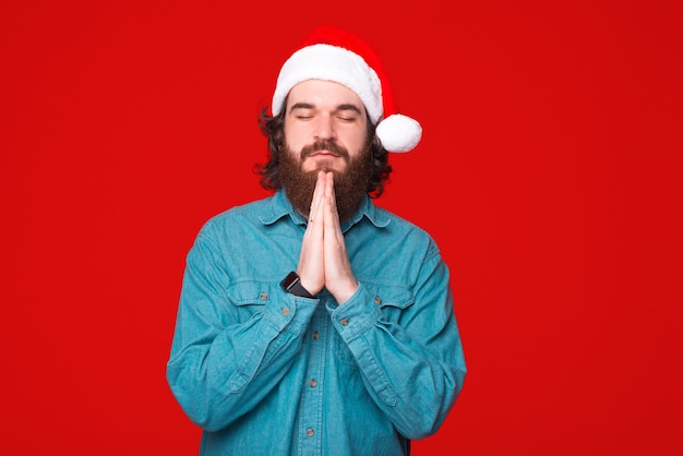 Giovane uomo barbuto che indossa il cappello di Natale sta tenendo le mani in preghiera su sfondo rosso