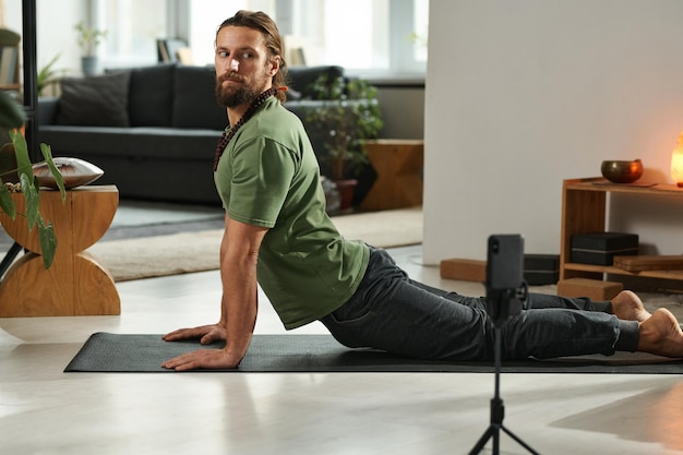 Giovane uomo barbuto che riprende esercizi di yoga sullo smartphone in piedi sul treppiede di fronte a lui che sta facendo
