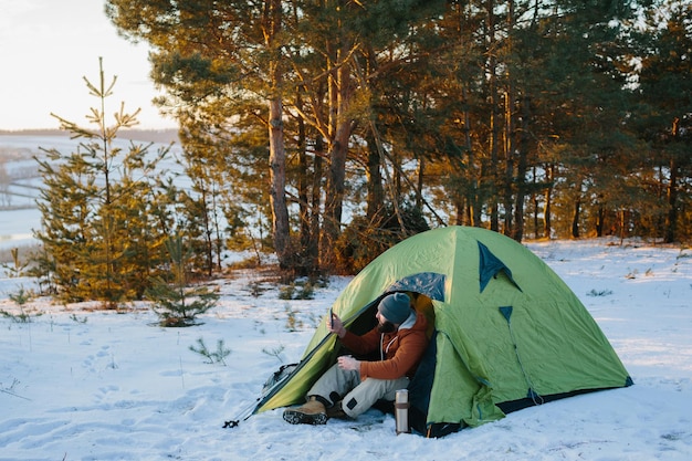 Giovane uomo barbuto che sorride bevendo caffè e prendendo selfie nelle montagne invernali vicino alla tenda