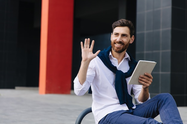 Giovane uomo barbuto che sorride e saluta qualcuno mentre è seduto su una panchina con tavoletta digitale. Concetto di persone, lavoro e tecnologia.