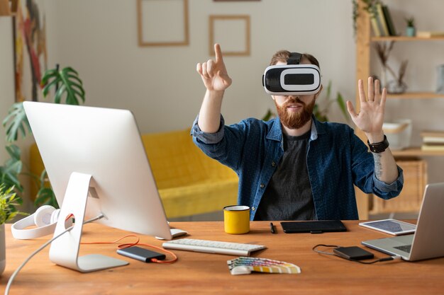 Giovane uomo barbuto che tocca lo schermo invisibile mentre si lavora sul design virtuale in cuffie da realtà virtuale