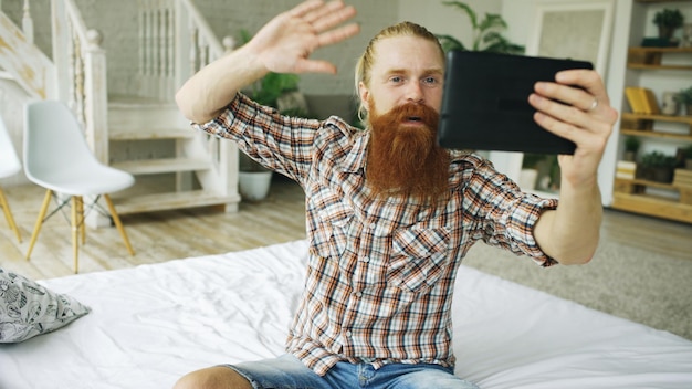 Giovane uomo barbuto che utilizza un computer tablet con chat video seduto a letto a casa