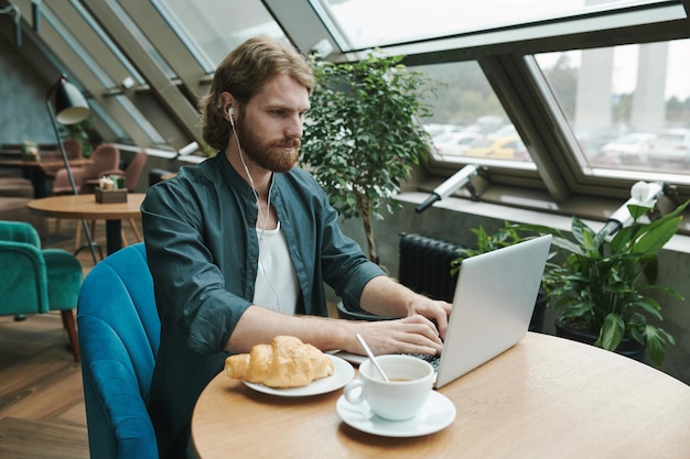 Giovane uomo barbuto concentrato in camicia casual che beve caffè e mangia croissant mentre controlla s