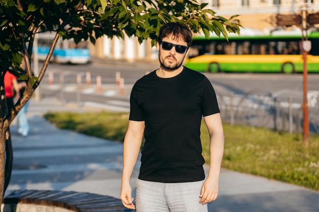 Giovane uomo barbuto elegante in una maglietta nera e occhiali da sole Foto di strada