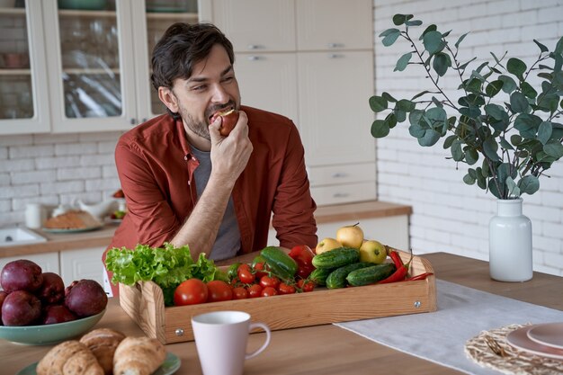 Giovane uomo barbuto felice in abiti casual che mangia mela rossa e sorride mentre sta in piedi nel moderno