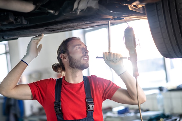 Giovane uomo barbuto in guanti protettivi in piedi sotto l'auto controllando le sue condizioni tecniche