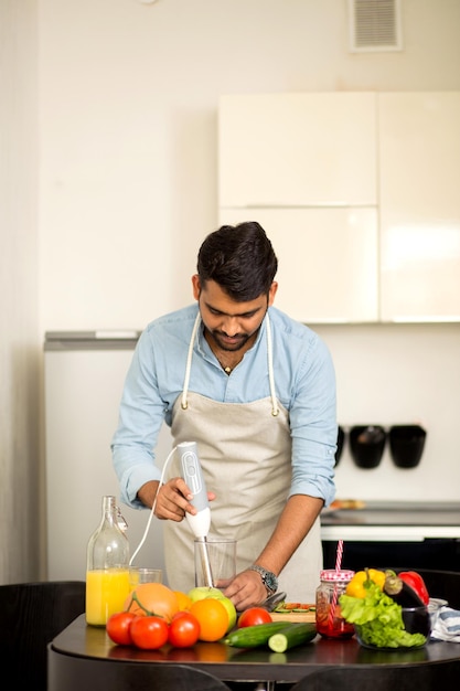 Giovane uomo barbuto indiano che indossa camicia e grembiule che cucina succo di vitamina fresco, frullato, cocktail con verdure crude e frutta nel frullatore in cucina. Cibo sano, dieta, concetto di pasto.