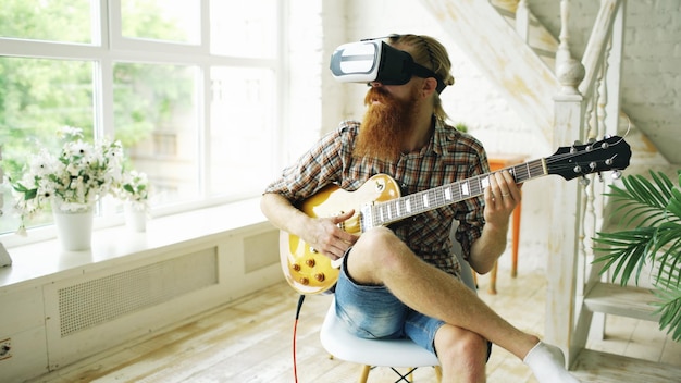 Giovane uomo barbuto seduto su una sedia che impara a suonare la chitarra utilizzando l'auricolare VR 360 a casa