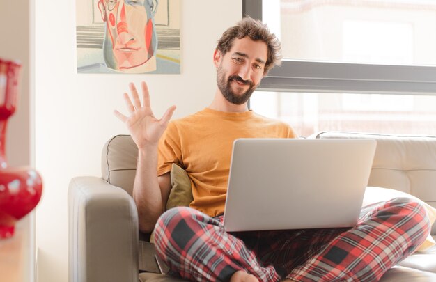 Giovane uomo barbuto sorridente e guardando amichevole che mostra il numero cinque o quinto con la mano in avanti il conto alla rovescia e seduto con un computer portatile