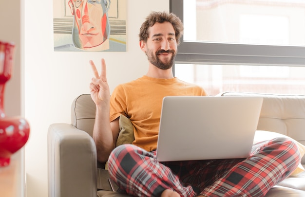 Giovane uomo barbuto sorridente e guardando amichevole che mostra il numero due o secondi con la mano in avanti il conto alla rovescia e seduto con un computer portatile