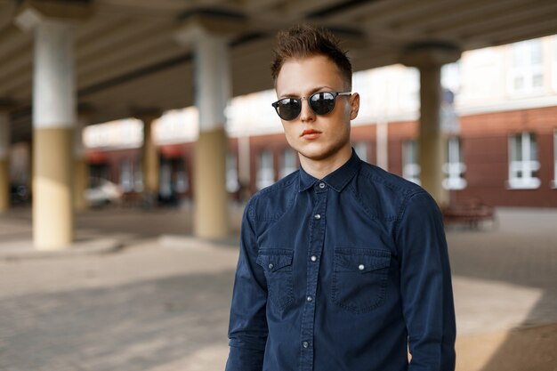 Giovane uomo bello alla moda in occhiali da sole e una camicia blu in una giornata di sole sulla strada