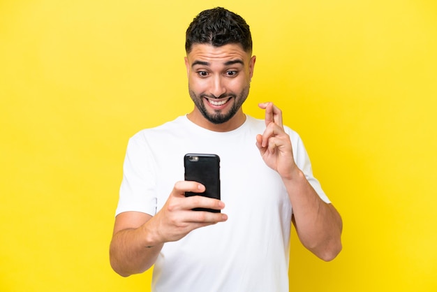 Giovane uomo bello arabo isolato su sfondo giallo utilizzando il telefono cellulare con le dita che si incrociano