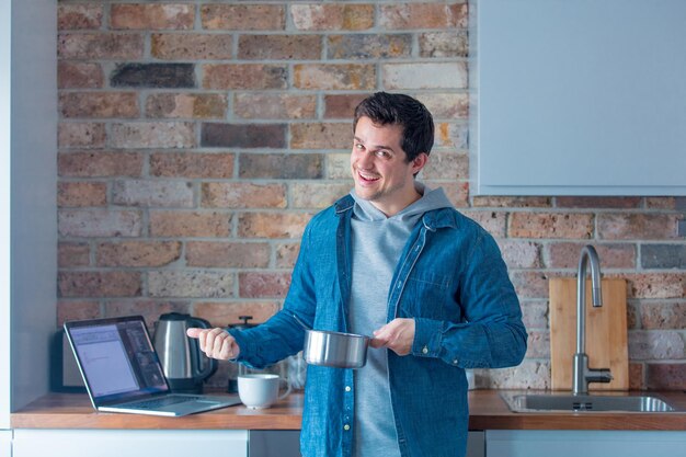 Giovane uomo bello che cucina in cucina utilizzando internet tramite computer portatile