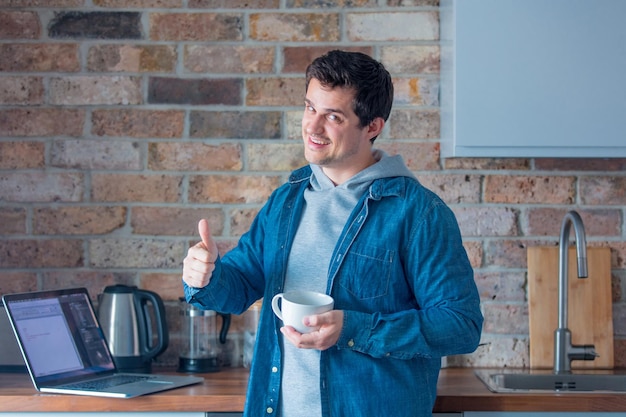 Giovane uomo bello che cucina in cucina utilizzando internet tramite computer portatile