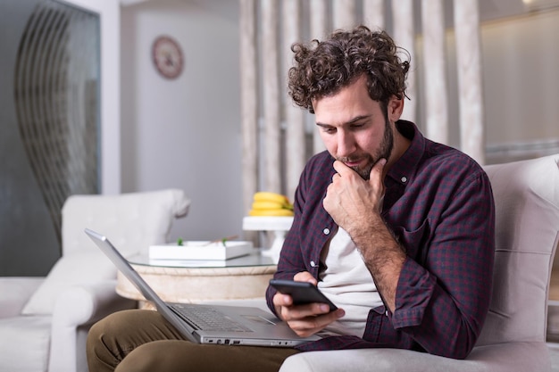 Giovane uomo bello che fa una videochiamata con gli amici mentre è seduto sul divano della sua casa moderna. Concetto di uomini d'affari felici. Uomo d'affari a casa che controlla le statistiche su telefono cellulare e laptop