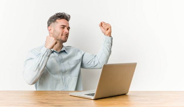 Giovane uomo bello che lavora con il suo computer portatile che alza pugno dopo una vittoria, concetto del vincitore.