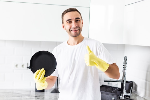 Giovane uomo bello che mostra i pollici in su lavare i piatti in cucina