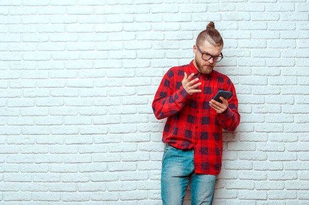 Giovane uomo bello che per mezzo del telefono cellulare mentre appoggiandosi muro di mattoni