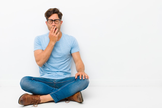 Giovane uomo bello che sbadiglia pigramente al mattino presto, svegliarsi e sembrare assonnato, stanco e annoiato seduto sul pavimento