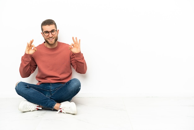 Giovane uomo bello che si siede sul pavimento che mostra un segno giusto con le dita