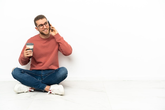 Giovane uomo bello che si siede sul pavimento che tiene il caffè da portare via e un cellulare