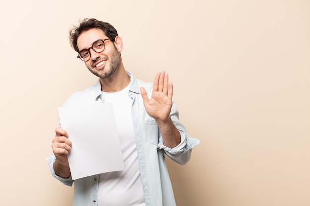 Giovane uomo bello che sorride allegramente e allegramente, agitando la mano, dandoti il benvenuto e salutandoti o salutandoti