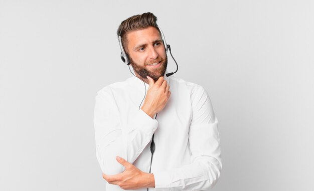 Giovane uomo bello che sorride con un'espressione felice e sicura con la mano sul mento. concetto di telemarketing