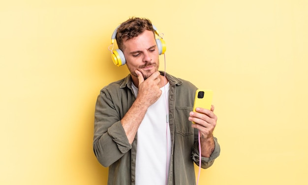 Giovane uomo bello che sorride con un'espressione felice e sicura con la mano sul mento cuffie e concetto di smartphone