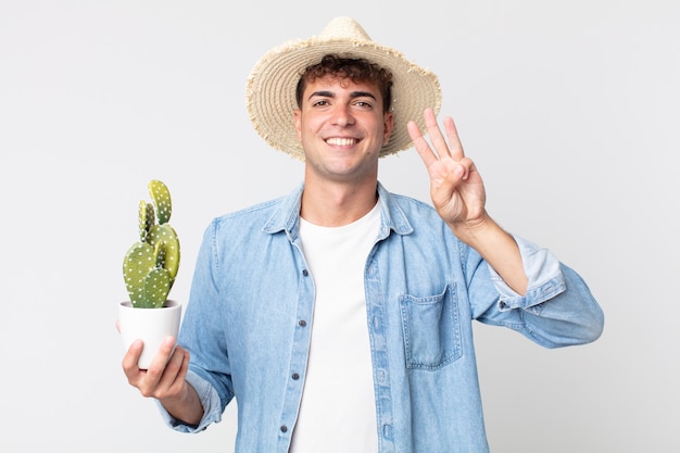 Giovane uomo bello che sorride e sembra amichevole, mostrando il numero tre. contadino con in mano un cactus decorativo