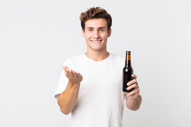 Giovane uomo bello che sorride felicemente con amichevole e offrendo e mostrando un concetto e tenendo in mano una bottiglia di birra