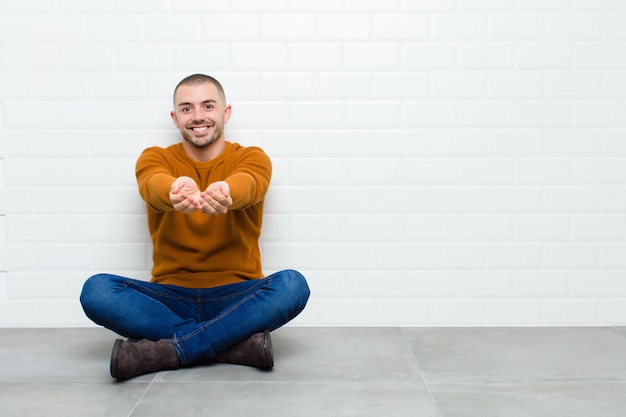 Giovane uomo bello che sorride felicemente con lo sguardo amichevole, sicuro, positivo, che offre e che mostra un oggetto o un concetto che si siede sul pavimento