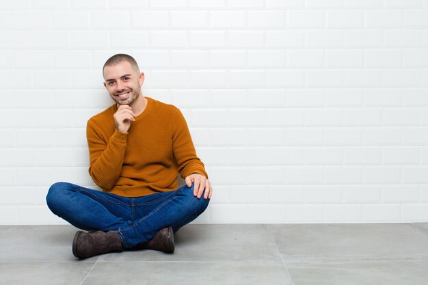 Giovane uomo bello che sorride, godendosi la vita, sentendosi felice, amichevole, soddisfatto e spensierato con la mano sul mento che si siede sul pavimento