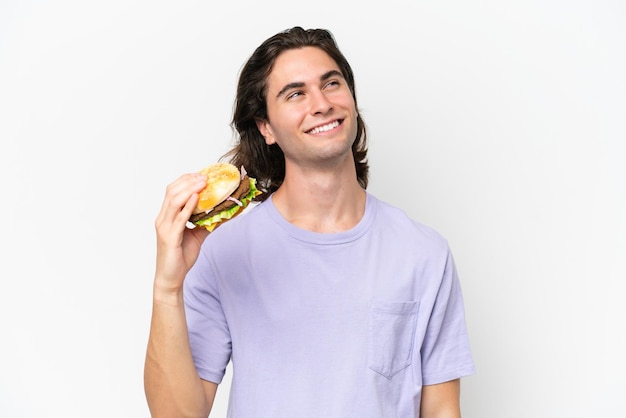 Giovane uomo bello che tiene un hamburger isolato su sfondo bianco alzando lo sguardo mentre sorride