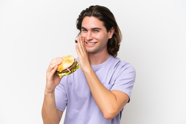 Giovane uomo bello che tiene un hamburger isolato su sfondo bianco sussurrando qualcosa