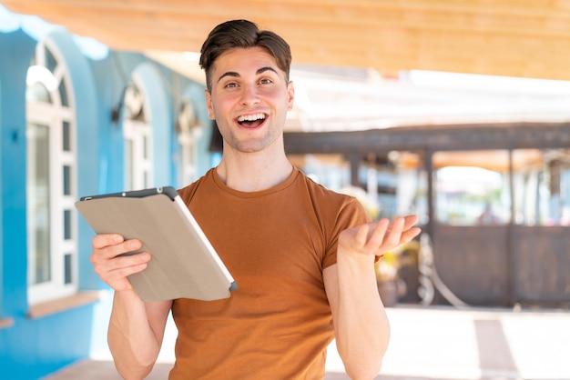 Giovane uomo bello che tiene un tablet all'aperto con un'espressione facciale scioccata