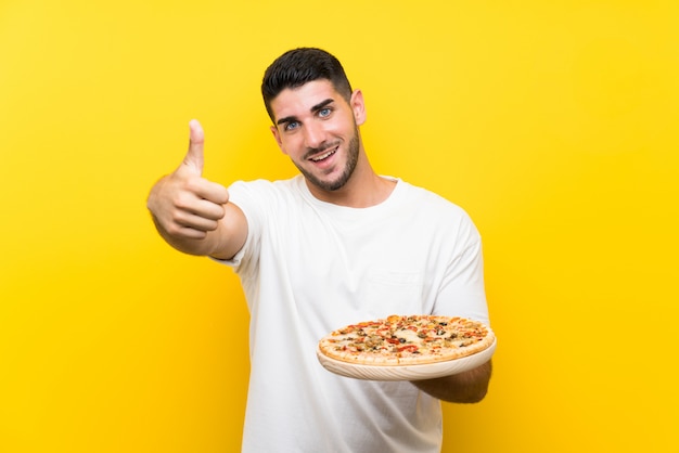 Giovane uomo bello che tiene una pizza sopra la parete gialla con i pollici in su perché è successo qualcosa di buono