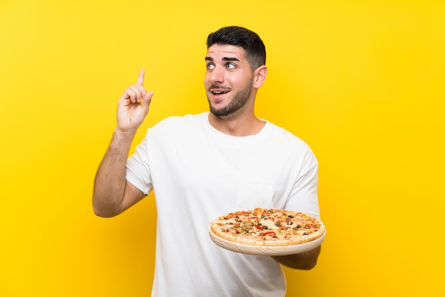 Giovane uomo bello che tiene una pizza sopra la parete gialla isolata che intende realizzare la soluzione mentre sollevando un dito su