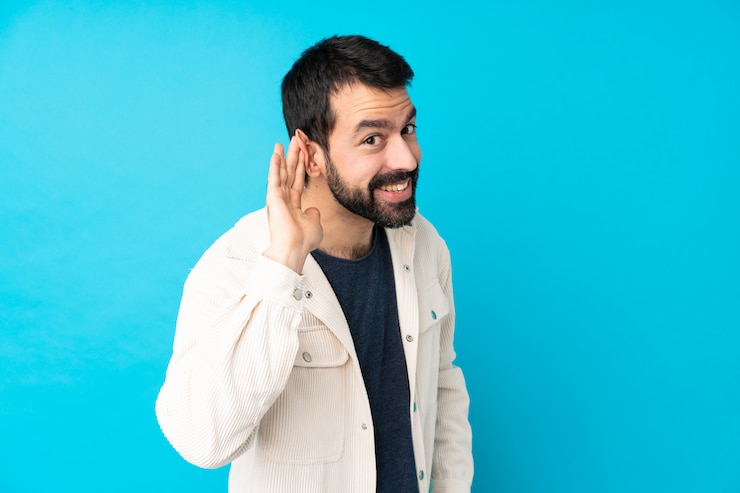 Foto giovane uomo bello con giacca di velluto bianco ascoltando qualcosa mettendo la mano sull'orecchio