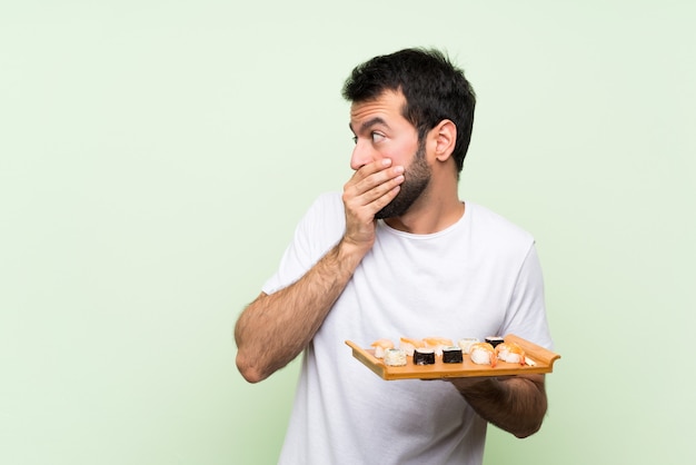 Giovane uomo bello con i sushi sopra la bocca verde isolata del rivestimento murale e guardare al lato