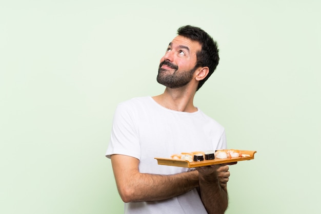 Giovane uomo bello con i sushi sopra la parete verde che osserva in su mentre sorridendo