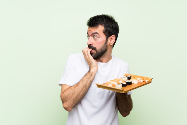 Giovane uomo bello con i sushi sopra la parete verde isolata nervosa e spaventata mettendo le mani alla bocca