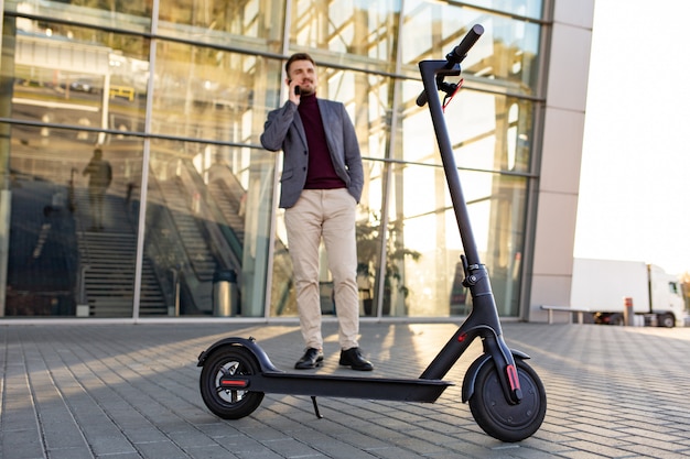 Giovane uomo bello con il motorino elettronico che sta sul marciapiede vicino all'aeroporto sul tramonto e che parla sullo smartphone