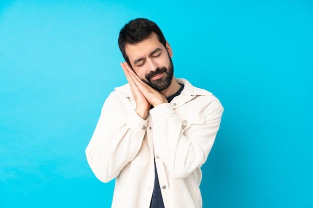 Giovane uomo bello con il rivestimento bianco del velluto a coste sopra la parete blu isolata che fa gesto di sonno nell'espressione dorable