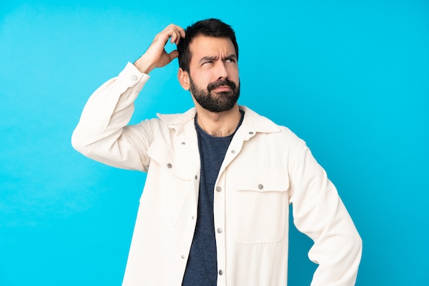 Giovane uomo bello con il rivestimento bianco del velluto a coste sopra la parete blu isolata che ha dubbi mentre graffiando testa