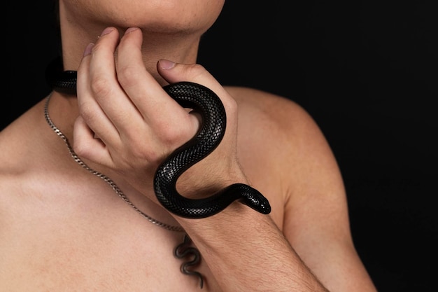 Giovane uomo bello con il torso nudo con un serpente nero che striscia intorno al collo isolato su sfondo nero