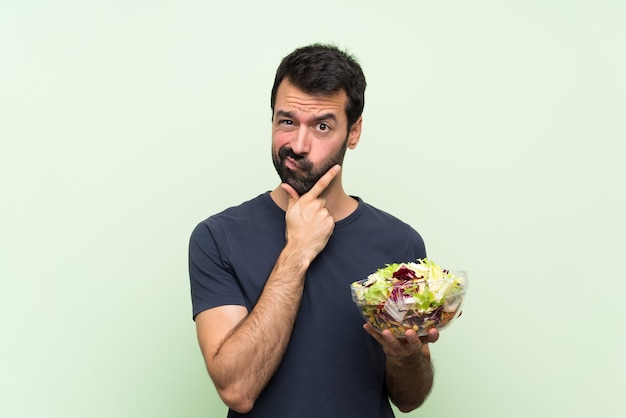 Giovane uomo bello con insalata sopra il pensiero verde isolato della parete