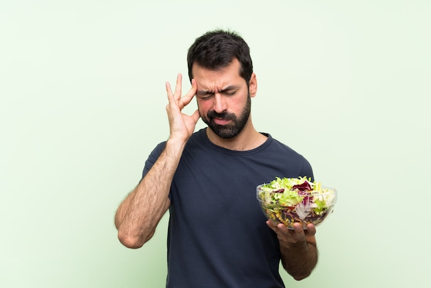 Giovane uomo bello con insalata sopra la parete verde isolata con l'emicrania